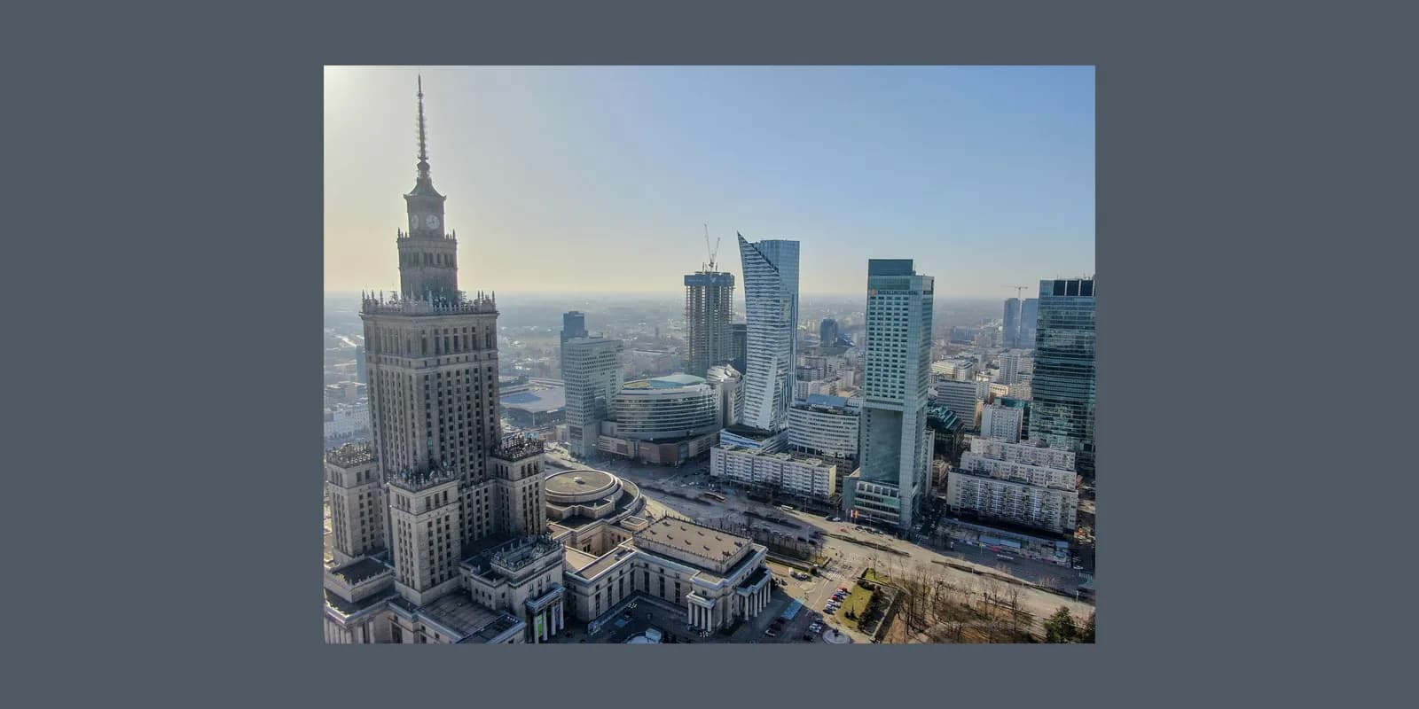 An aerial view of the Warsaw city skyline with the Palace of Culture and Science in the foreground, surrounded by modern skyscrapers under a bright sky.