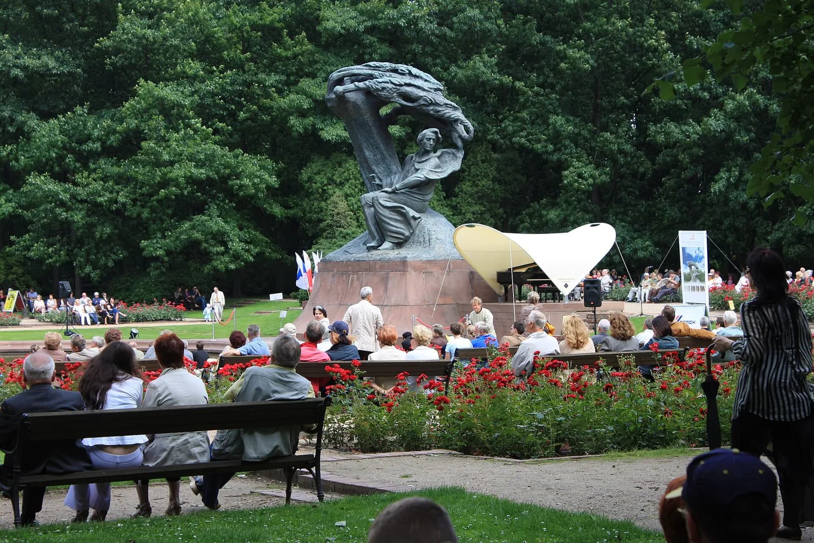 A large bronze statue of Frédéric Chopin under a weeping willow tree, overlooking an audience seated on benches in a garden with red roses, watching a musician perform on a stage under a white tent.