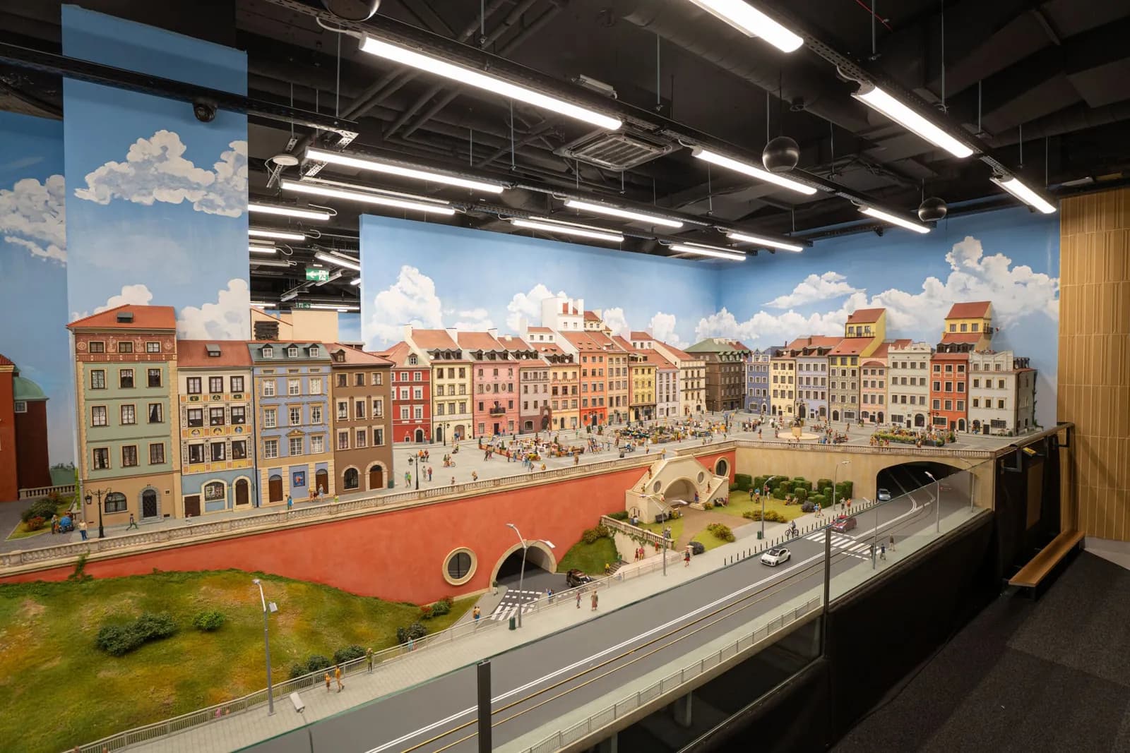 A detailed miniature city scene features colorful buildings, a bustling town square with tiny people, and a road with miniature cars and tunnels, set against a backdrop of a blue sky with white clouds.